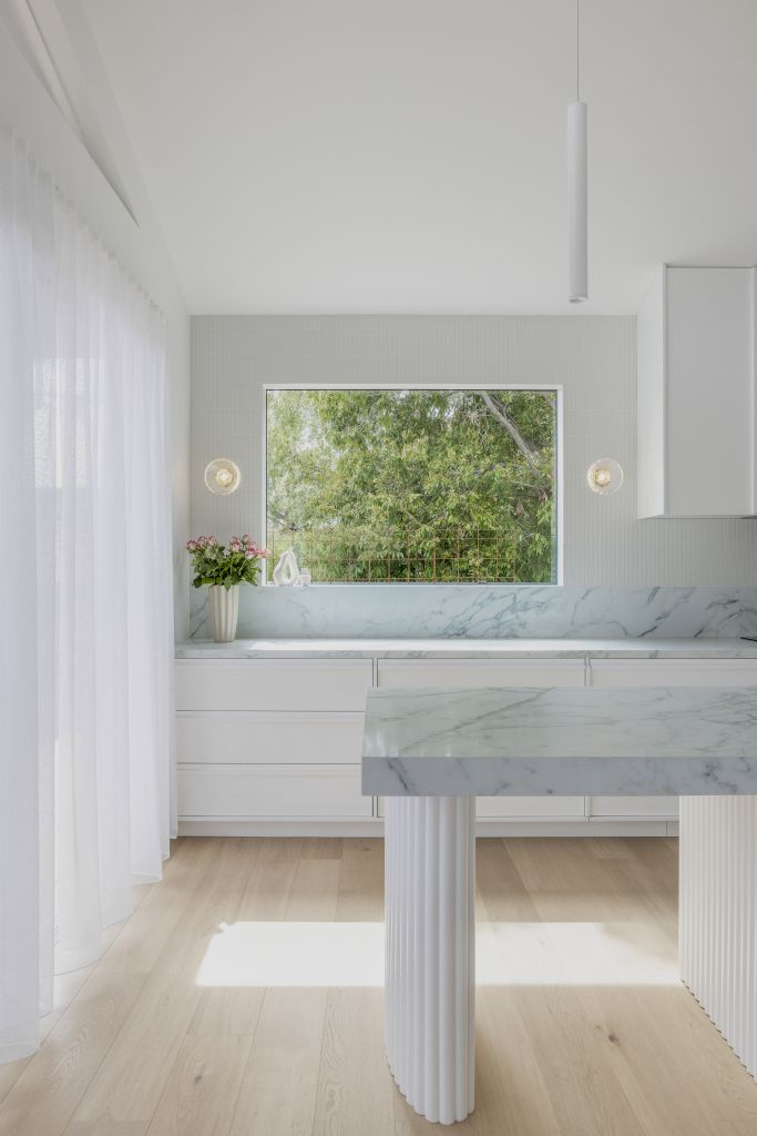 Kitchen nook with marble benchtop and splashback, sheer curtains and window framed by greenery