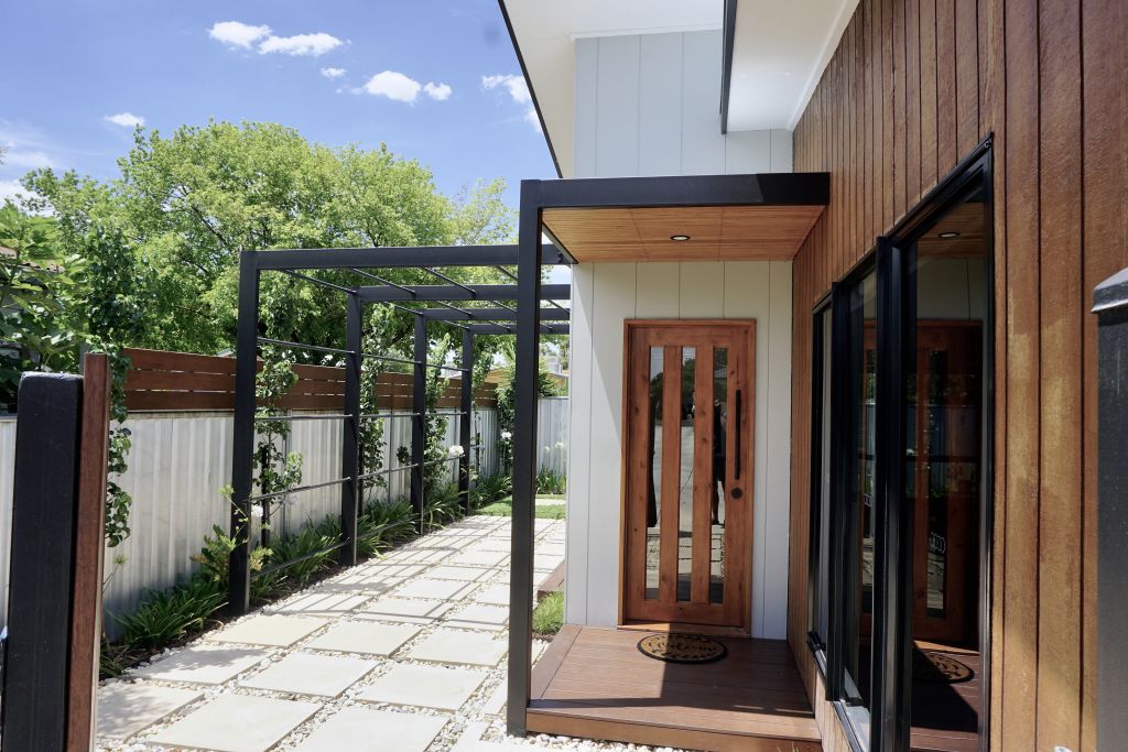 Front entrance with timber door, black-framed pergola, stone paver pathway and timber-clad facade