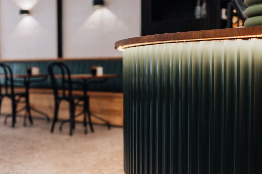 Close-up of curved green fluted bar counter with timber top and ambient lighting, with banquette seating in background