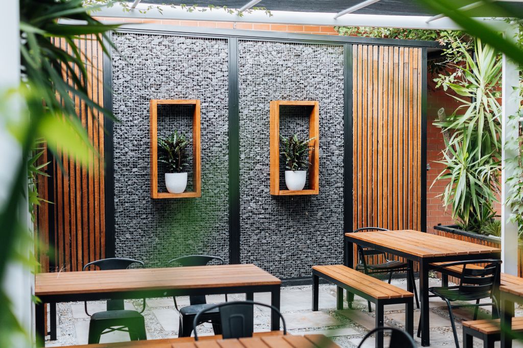 Outdoor courtyard seating area with timber tables, gabion stone wall, framed planters and surrounding greenery